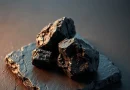 Bongkahan batu bara hitam bertekstur tajam di atas permukaan batu gelap dengan pencahayaan editorial.